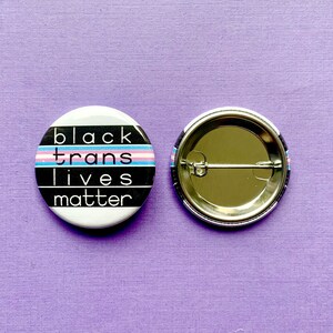 Puede incluir: Bot&oacute;n blanco y negro con el texto "black trans lives matter" y una bandera del orgullo transg&eacute;nero. La parte posterior del bot&oacute;n es plateada.