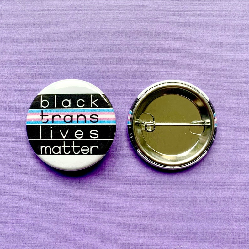 Puede incluir: Bot&oacute;n blanco y negro con el texto "black trans lives matter" y una bandera del orgullo transg&eacute;nero. La parte posterior del bot&oacute;n es plateada.