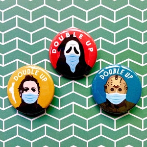 May include: Three round buttons with a yellow, red, and blue background. Each button features a different horror movie character wearing a face mask. The text "DOUBLE UP" is printed on each button.