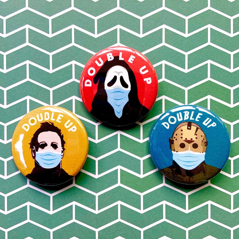 May include: Three round buttons with a yellow, red, and blue background. Each button features a different horror movie character wearing a face mask. The text "DOUBLE UP" is printed on each button.