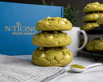May include: Three green cookies with white chocolate chips stacked on top of each other. A blue box with the text "N-Ticing Share Something" is in the background. A white mug and a spoon with green powder are also visible.