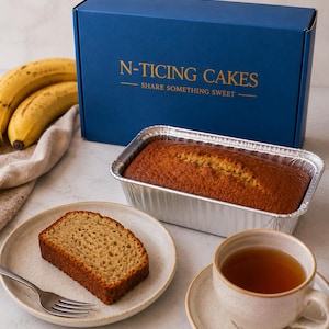 May include: A photograph of a freshly baked banana loaf in a foil tin, with a slice on a plate and a cup of tea. A blue box with gold lettering reading "N-Ticing Cakes - Share Something Sweet" is in the background. Ripe bananas are also visible.