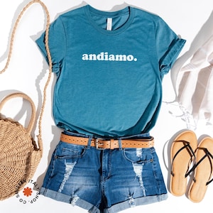 May include: A teal blue t-shirt with the text "andiamo." printed in white on the front. The shirt is paired with denim shorts, a brown belt, and brown sandals.