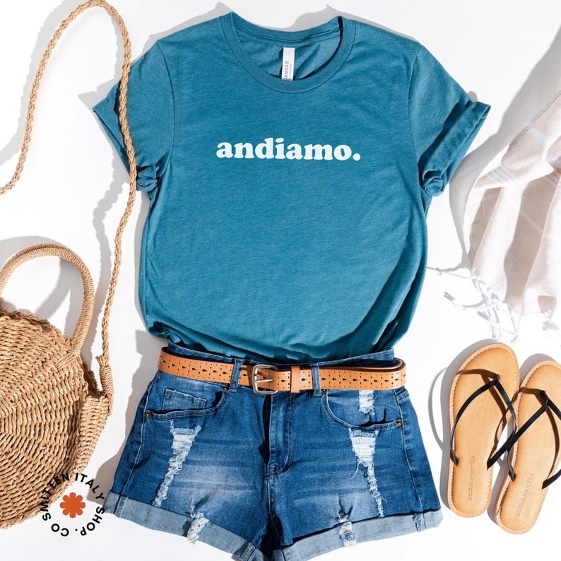 May include: A teal blue t-shirt with the text "andiamo." printed in white on the front. The shirt is paired with denim shorts, a brown belt, and brown sandals.