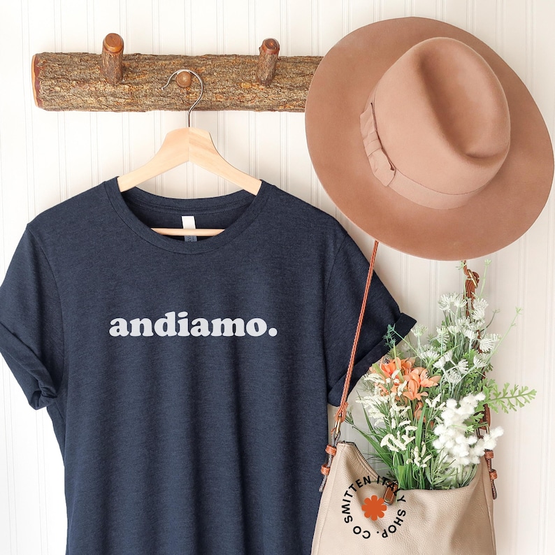 May include: A navy blue t-shirt with the text "andiamo." printed in white on the front. The shirt is hanging on a wooden hanger against a white wall.