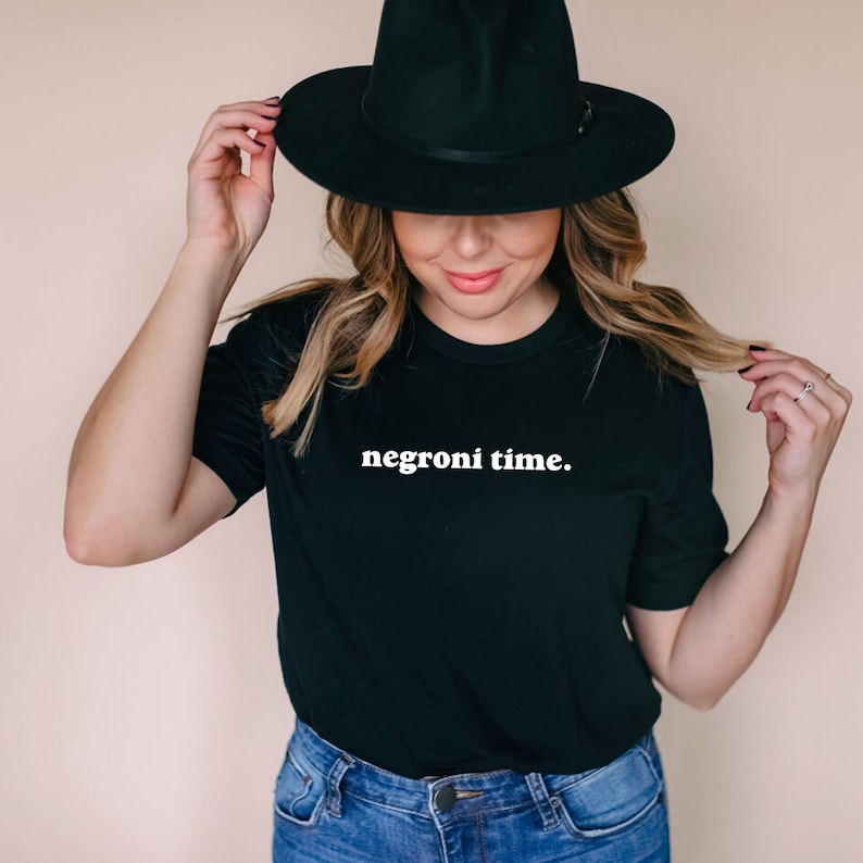 Może przedstawiać: Kobieta w czarnym t-shircie z napisem "negroni time." w białych literach. Ma na sobie r&oacute;wnież czarny kapelusz fedora i niebieskie dżinsy.