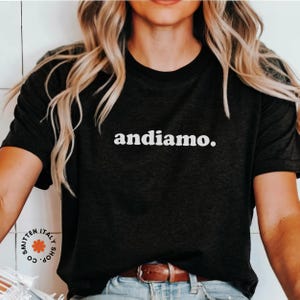 May include: A black t-shirt with the text "andiamo." printed in white on the front. The shirt is being worn by a person with long blonde hair.