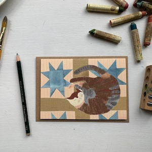 May include: A hand-painted card featuring a cat sleeping on a blue and brown patchwork quilt. The quilt has a star pattern.