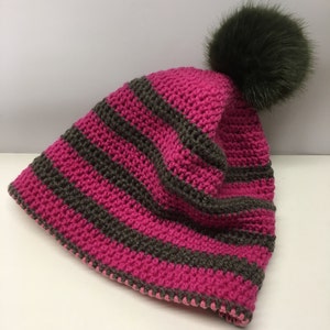 Könnte beinhalten: Eine gestrickte Mütze in rosa und braunen Streifen mit einem dunkelbraunen Kunstfellbommel oben.
