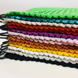 Könnte beinhalten: Ein Stapel bunter, handgehäkelter Waschlappen in verschiedenen Farbtönen, darunter Grün, Orange, Lila und Gelb. Jeder Waschlappen ist mit einem schwarzen Metallring versehen. Der Stapel befindet sich auf einer weißen Oberfläche.