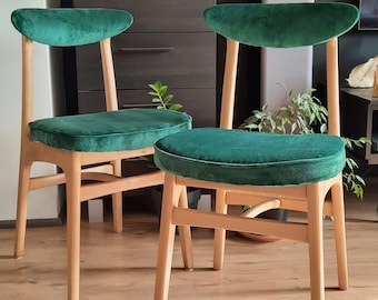 May include: Two green velvet dining chairs with wooden legs and a simple design. The chairs are on a light wood floor.