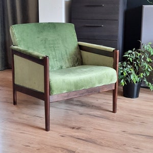 May include: A green velvet armchair with dark wood frame and legs. The chair is sitting on a wooden floor in a room with a dresser and plants.