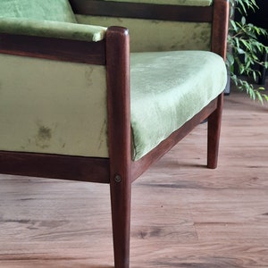 May include: A green velvet armchair with dark wood legs and a wooden frame. The chair is sitting on a wooden floor.