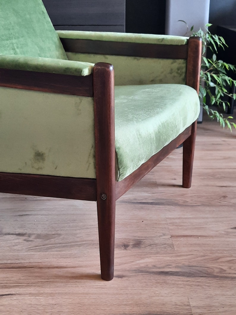 May include: A green velvet armchair with dark wood legs and a wooden frame. The chair is sitting on a wooden floor.