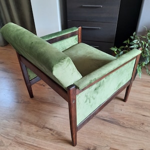 May include: A green velvet armchair with a wooden frame. The chair has a high back and a wide seat. The chair is sitting on a wooden floor.