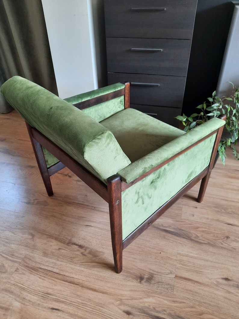 May include: A green velvet armchair with a wooden frame. The chair has a high back and a wide seat. The chair is sitting on a wooden floor.