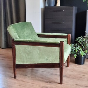 May include: A green velvet armchair with a dark wood frame. The chair has a low back and wide arms. The chair is sitting on a wooden floor.
