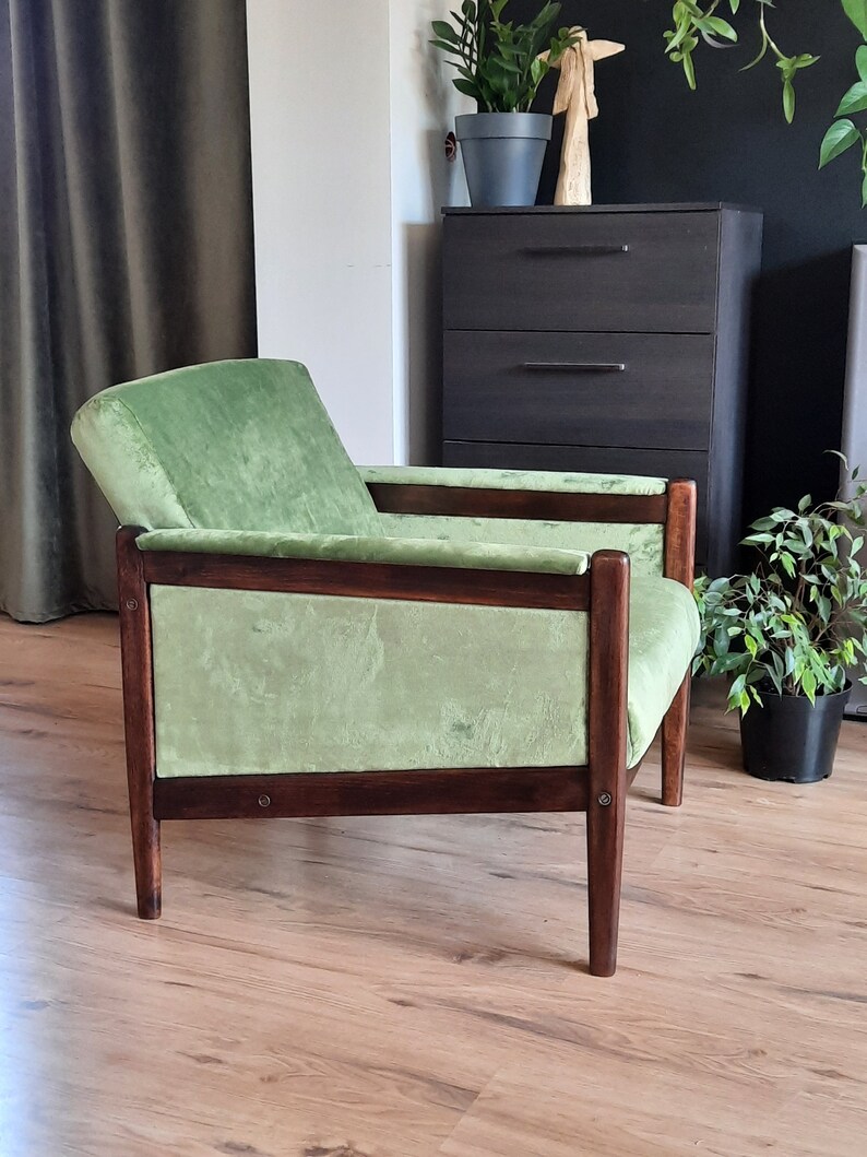 May include: A green velvet armchair with a dark wood frame. The chair has a low back and wide arms. The chair is sitting on a wooden floor.