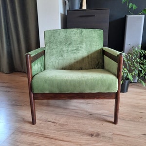 May include: A green velvet armchair with dark wood legs and arms. The chair is sitting on a wooden floor.