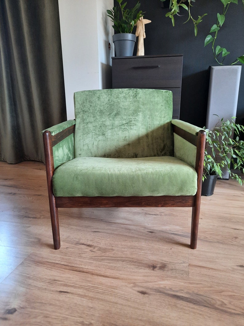 May include: A green velvet armchair with dark wood legs and arms. The chair is sitting on a wooden floor.