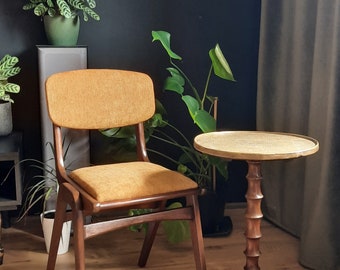 May include: A mid-century modern chair with a mustard-colored seat and backrest. The chair has a wooden frame and legs. A small side table with a brass top and a bamboo-style base is next to the chair. The room has plants and a dark wall.