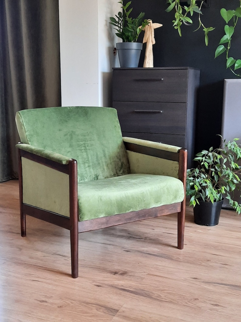 May include: A green velvet armchair with dark wood frame and legs. The chair is sitting on a wooden floor in a room with a dresser and plants.
