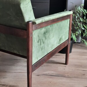 May include: A green velvet armchair with a dark wood frame. The chair has a high back and a wide seat. The chair is sitting on a wooden floor.