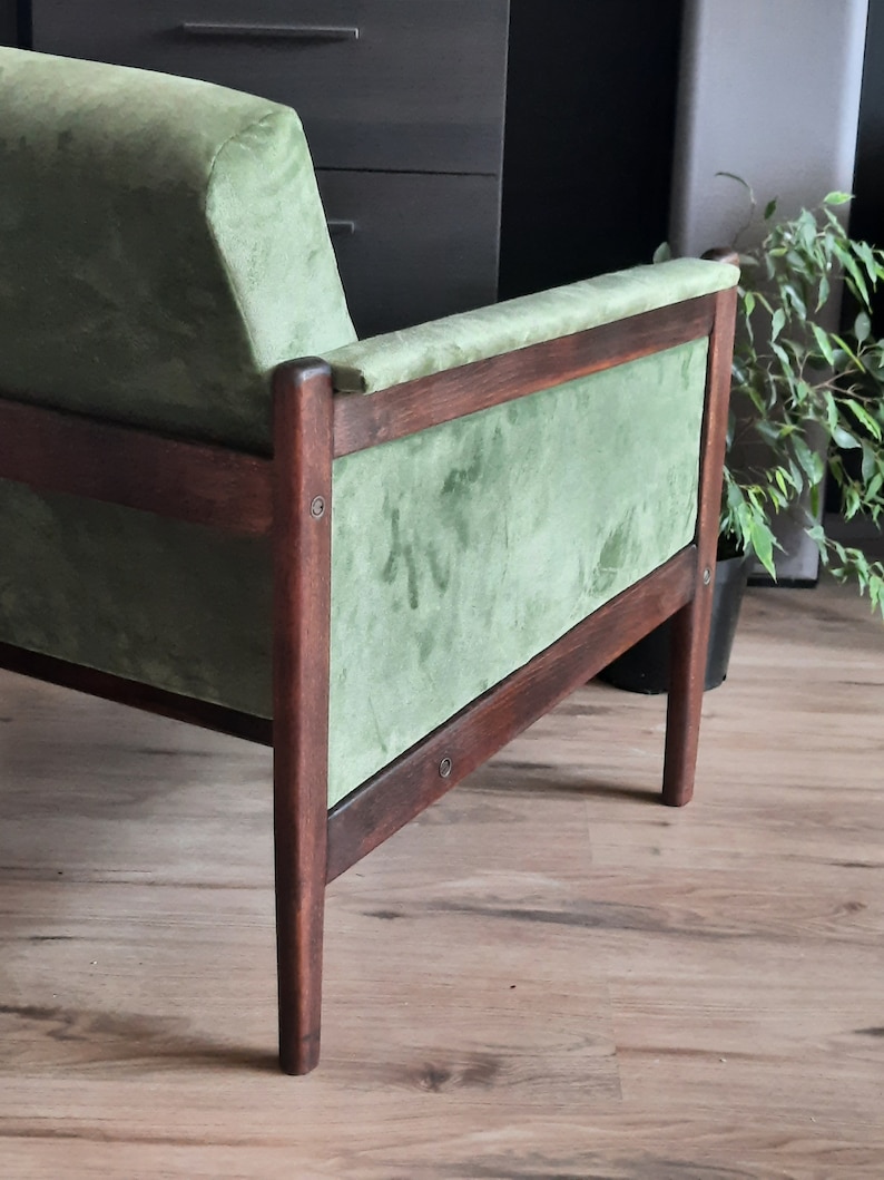 May include: A green velvet armchair with a dark wood frame. The chair has a high back and a wide seat. The chair is sitting on a wooden floor.
