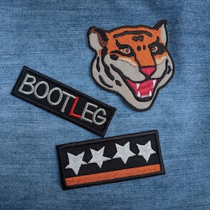 Könnte beinhalten: Drei gestickte Patches auf einem blauen Jeans-Hintergrund. Die Patches sind schwarz mit weißen Nähten. Ein Patch hat das Wort "BOOTLEG" in weißen Buchstaben. Ein anderer Patch hat fünf weiße Sterne. Der dritte Patch ist ein Tigerkopf mit orangefarbenen und schwarzen Streifen.