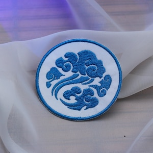 Puede incluir: Un parche bordado blanco y azul con un diseño estilizado de nubes y olas. El parche es circular y tiene un borde azul.