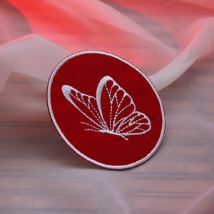 Op de afbeelding: Een rood geborduurd patch met een wit vlinderontwerp. De patch is rond en heeft een witte omtrek.