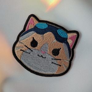 以下が含まれることがあります： 飛行士ゴーグルをかけた漫画の猫の顔の刺繍パッチ。猫はベージュ、グレー、ピンクのディテールがあり、黒いアウトラインがあります。ゴーグルは青色です。パッチは白い背景にあります。