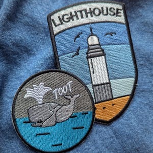 May include: Two embroidered patches on blue fabric. One is a shield shape with a lighthouse design and the word "LIGHTHOUSE". The other is a round patch with a whale and the word "TOOT".