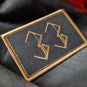 Peut inclure: Un écusson rectangulaire noir avec des bords brodés dorés et un motif géométrique doré. L'écusson est posé sur un fond sombre et texturé, avec une touche de rouge visible.