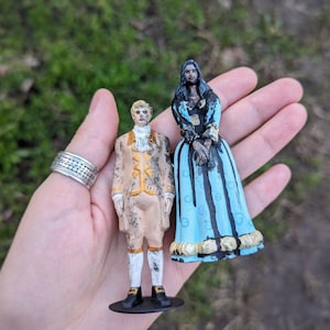 Puede incluir: Dos pequeñas figuritas de personas con ropa del siglo XVIII. La mujer lleva un vestido azul y negro con ribete de encaje blanco. El hombre lleva un traje marrón y dorado con ribete de encaje blanco. Ambas figuritas están de pie sobre bases negras.
