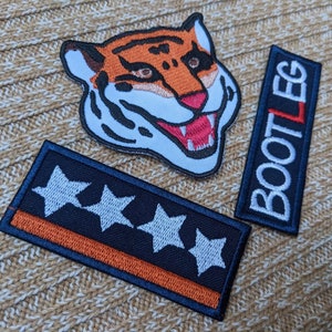 Puede incluir: Tres parches bordados: un parche azul con tres estrellas blancas y una franja naranja, un parche negro con la palabra "BOOTLEG" en blanco y un parche de cabeza de tigre con rayas naranjas y negras.