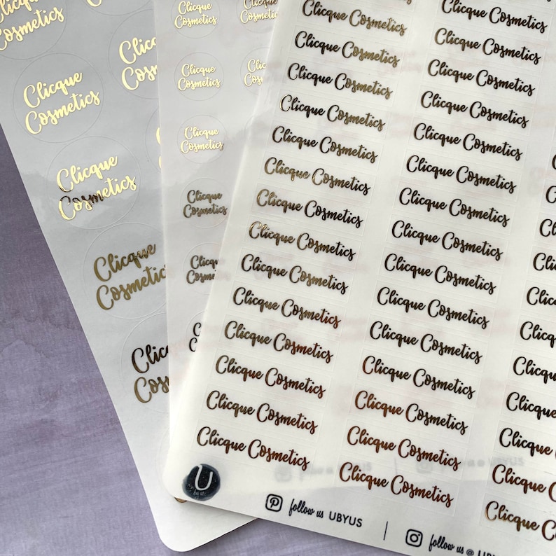 以下が含まれることがあります： 「Clicque Cosmetics」と書いてある金箔のラベルが貼られた透明なステッカー用紙です。ラベルは様々なサイズと形があり、円形のものもあります。