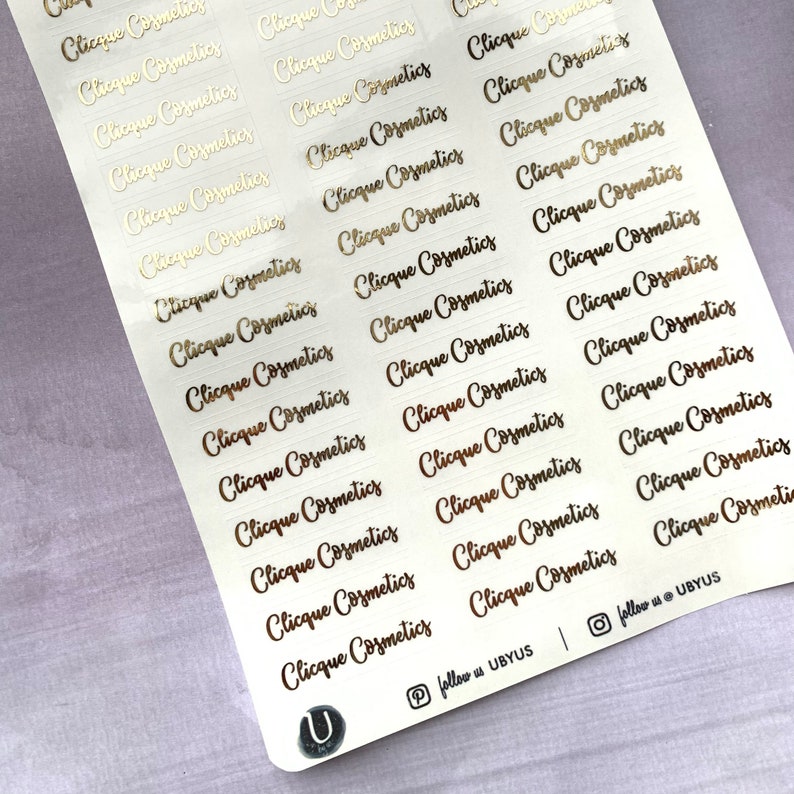 以下が含まれることがあります： 「Clicque Cosmetics」という文字が何度も繰り返された金箔ステッカーのシート。ステッカーは白い背景に貼られています。右下隅には「follow us @ UBYUS」という文字が印刷されています。