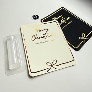 May include: Christmas gift tags in cream and black with gold foil lettering and bow accents. The cream tag reads "Merry Christmas" and "From: THE PHILLIPS FAMS." The black tag also says "Merry Christmas" and "From: THE PHILLIPS FAMILY."