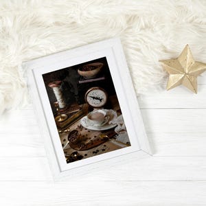 K&ouml;nnte beinhalten: Ein wei&szlig;er Bilderrahmen mit einem Vintage-Stillleben-Foto einer Tasse Kaffee, Kaffeebohnen, einer Waage und einer Garnrolle. Das Foto ist auf einer Holzoberfl&auml;che platziert.
