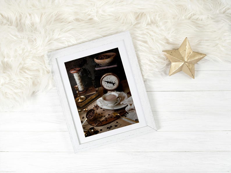K&ouml;nnte beinhalten: Ein wei&szlig;er Bilderrahmen mit einem Vintage-Stillleben-Foto einer Tasse Kaffee, Kaffeebohnen, einer Waage und einer Garnrolle. Das Foto ist auf einer Holzoberfl&auml;che platziert.