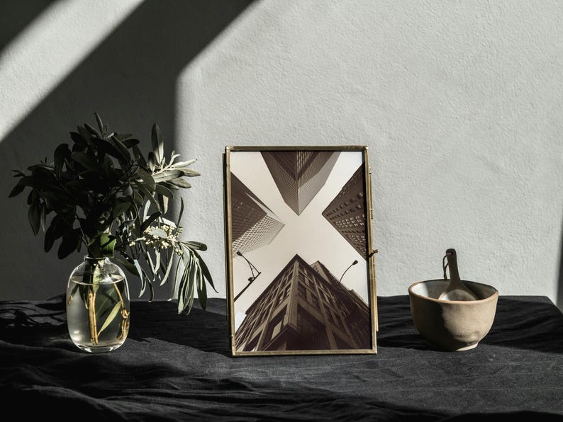 May include: A gold-toned picture frame with a black and white photograph of a city skyline. The frame is sitting on a black surface with a vase of greenery and a small bowl.