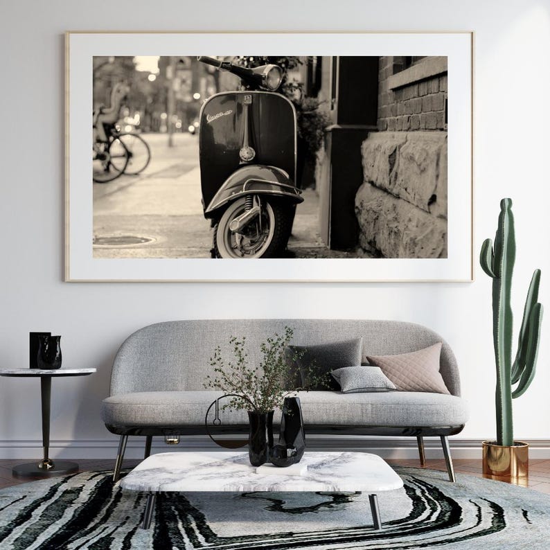 May include: A black and white photograph of a vintage Vespa scooter parked on a city street. The scooter is parked next to a brick building and there is a bicycle in the background.