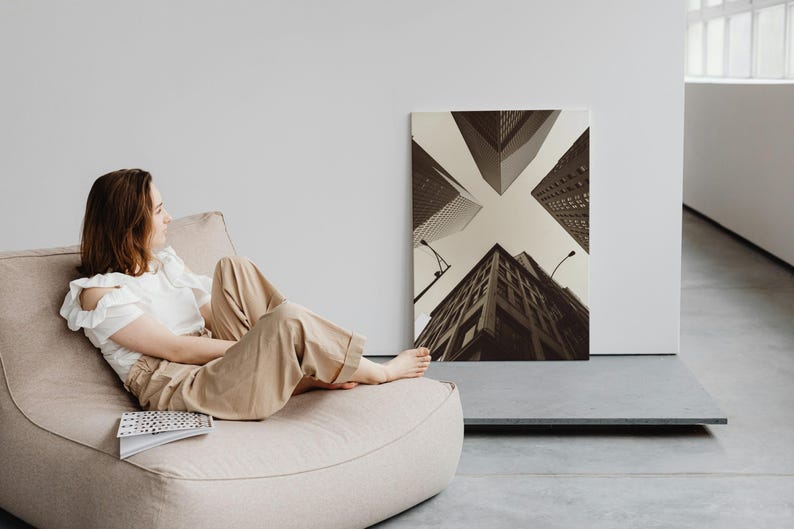May include: A woman relaxes in a beige beanbag chair, looking towards a black and white photograph of city buildings. The photograph is hanging on a white wall.