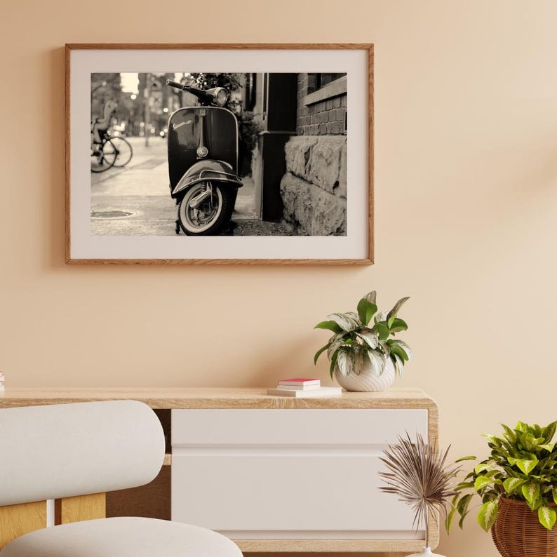 May include: A black and white photograph of a vintage Vespa scooter parked on a city street. The scooter is parked next to a brick building and there is a bicycle in the background.