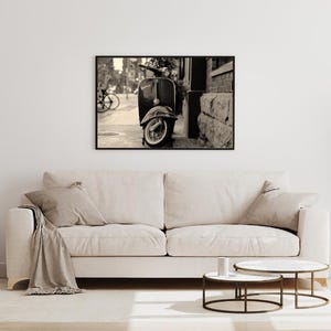 May include: Black and white photograph of a vintage scooter parked on a city street. The scooter is in focus, while the background is blurred.