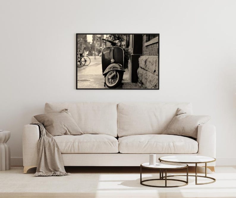 May include: Black and white photograph of a vintage scooter parked on a city street. The scooter is in focus, while the background is blurred.