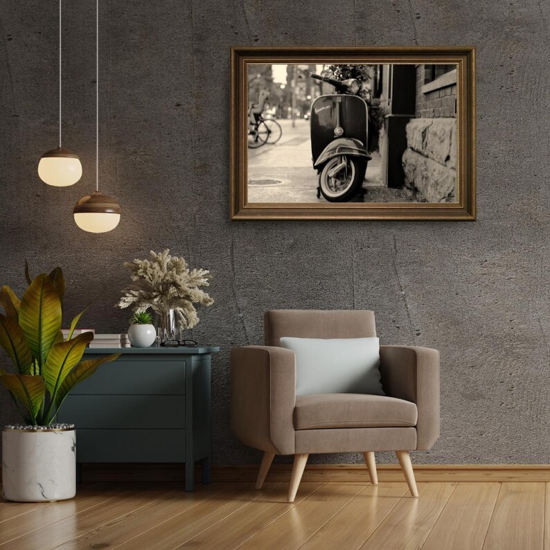 May include: A black and white photograph of a vintage scooter parked on a city street. The scooter is parked next to a brick building. The photo is framed in a brown wooden frame.