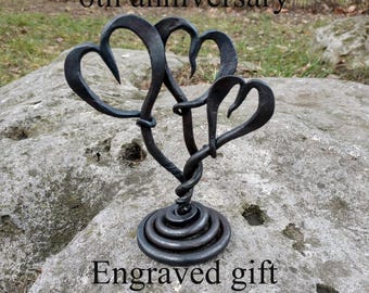 May include: A dark metal sculpture of intertwined hearts, with the text "6th anniversary" and "Engraved gift". The sculpture is set on a stone surface, with a spiral base and a twisted stem supporting the hearts.