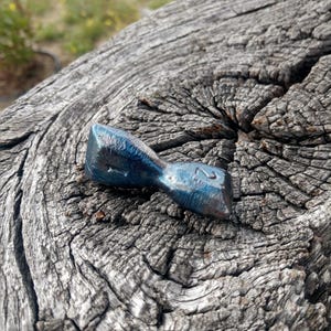 Puede incluir: Un dado de juego metálico azul, alargado y multifacético con una forma irregular distintiva. La superficie presenta un acabado oscuro e iridiscente con los números 1 y 2 grabados en sus caras angulares, mostrando un aspecto texturizado.